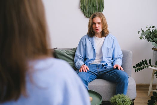 Concerned male client siting on sofa while having conversation about problems with faceless psychologist during psychotherapy appointment in light office